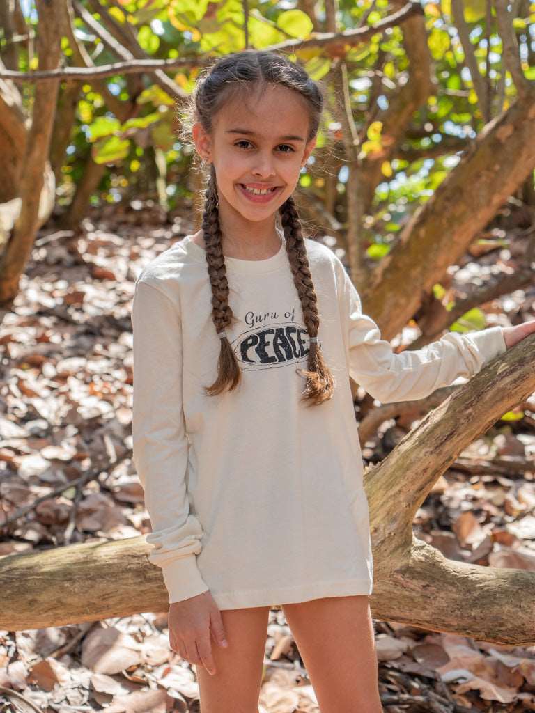 Young girl wearing a long sleeve shirt with the words 'Guru of Peace' sitting on a log in a forest setting