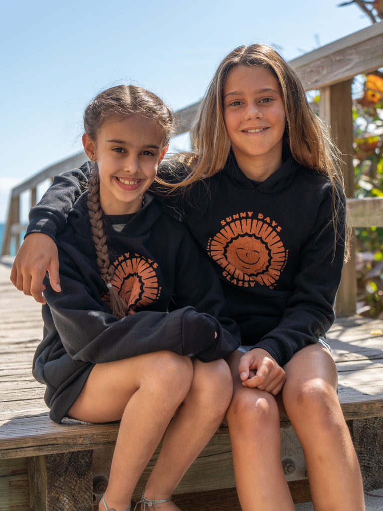 Two young girls wearing black hoodies with a sun design, sitting on a wooden bench outdoors.
