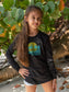 Young girl wearing a black long-sleeve shirt with a graphic design, standing on a beach.