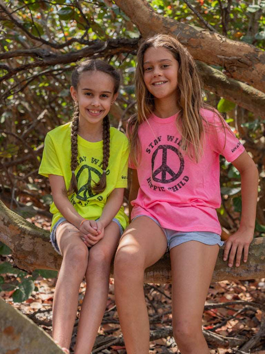 Two young girls sitting on a tree branch with peace signs on their shirts.