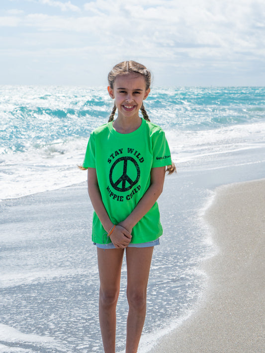 Young girl standing on a beach with a peace sign on her green shirt.